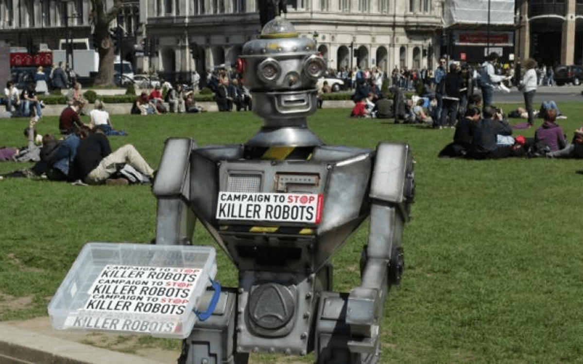 Robot standing in a crowded park wearing a sign reading "Campaigning to end killer robots" and handing out more signs reading the same