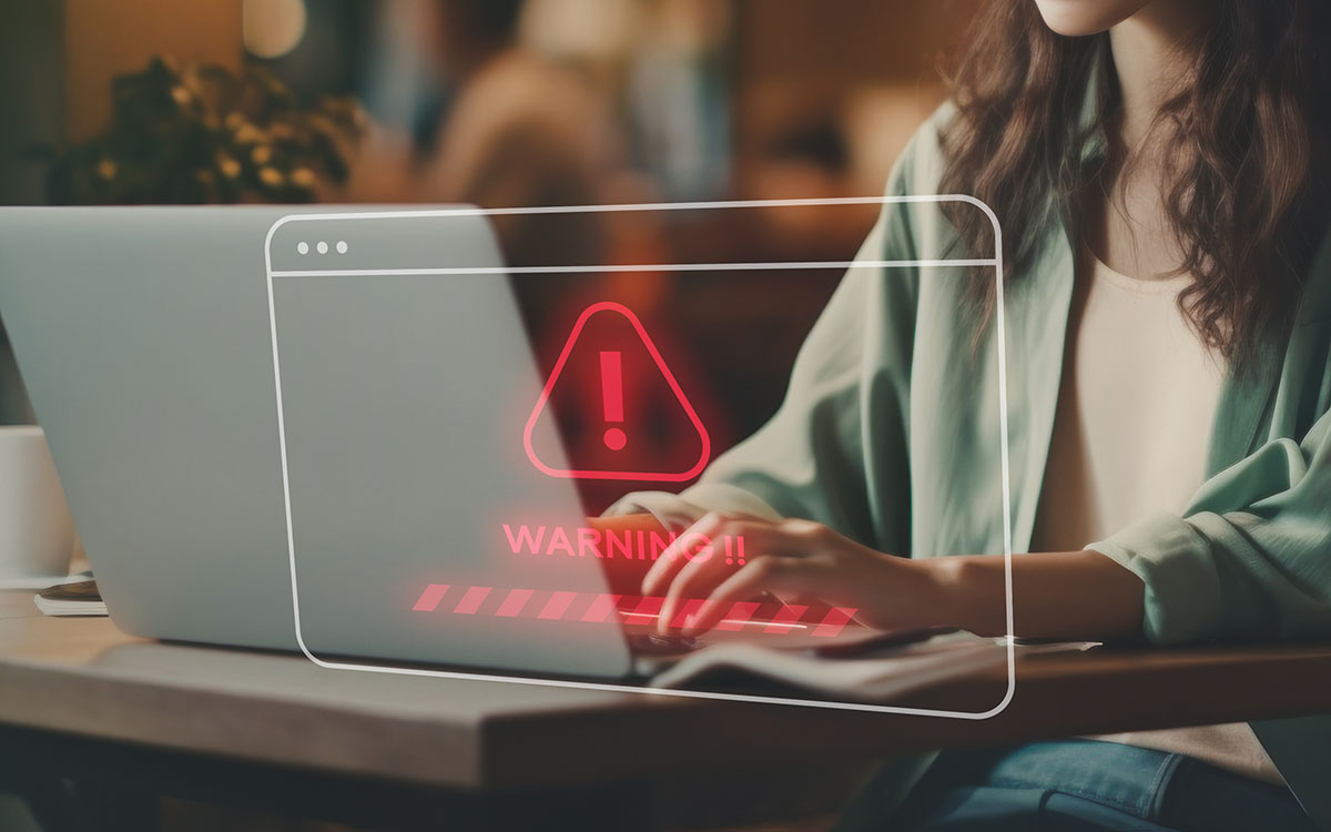 A woman using a laptop with a warning sign on it, indicating a potential credential stuffing attack.