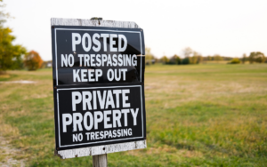 A sign on a wooden post reads "Posted No Trespassing Keep Out" and "Private Property No Trespassing" with a grassy field and trees in the background.