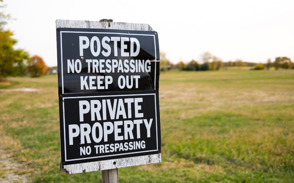 A sign on a wooden post reads "Posted No Trespassing Keep Out" and "Private Property No Trespassing" with a grassy field and trees in the background.